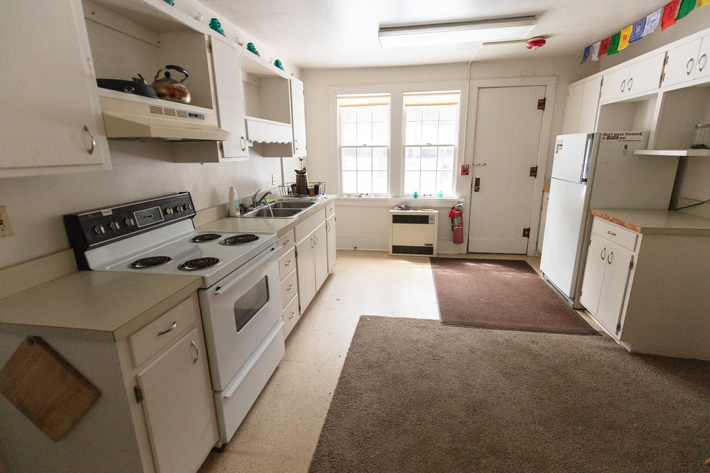 Canyon Mess Hall residential kitchen before renovation, with white cabinets, a range and sink along the left wall, and two rear windows.